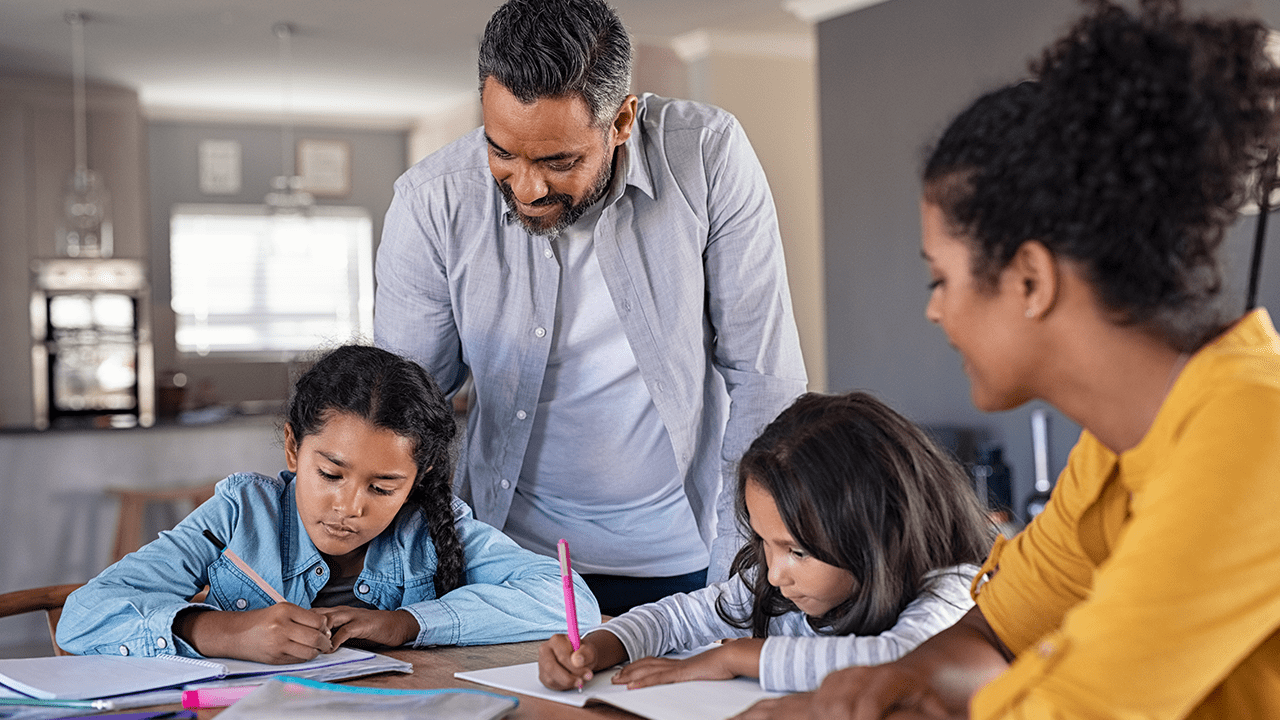 Parents sitting at the kitchen table and helping their two daughters with their homework.